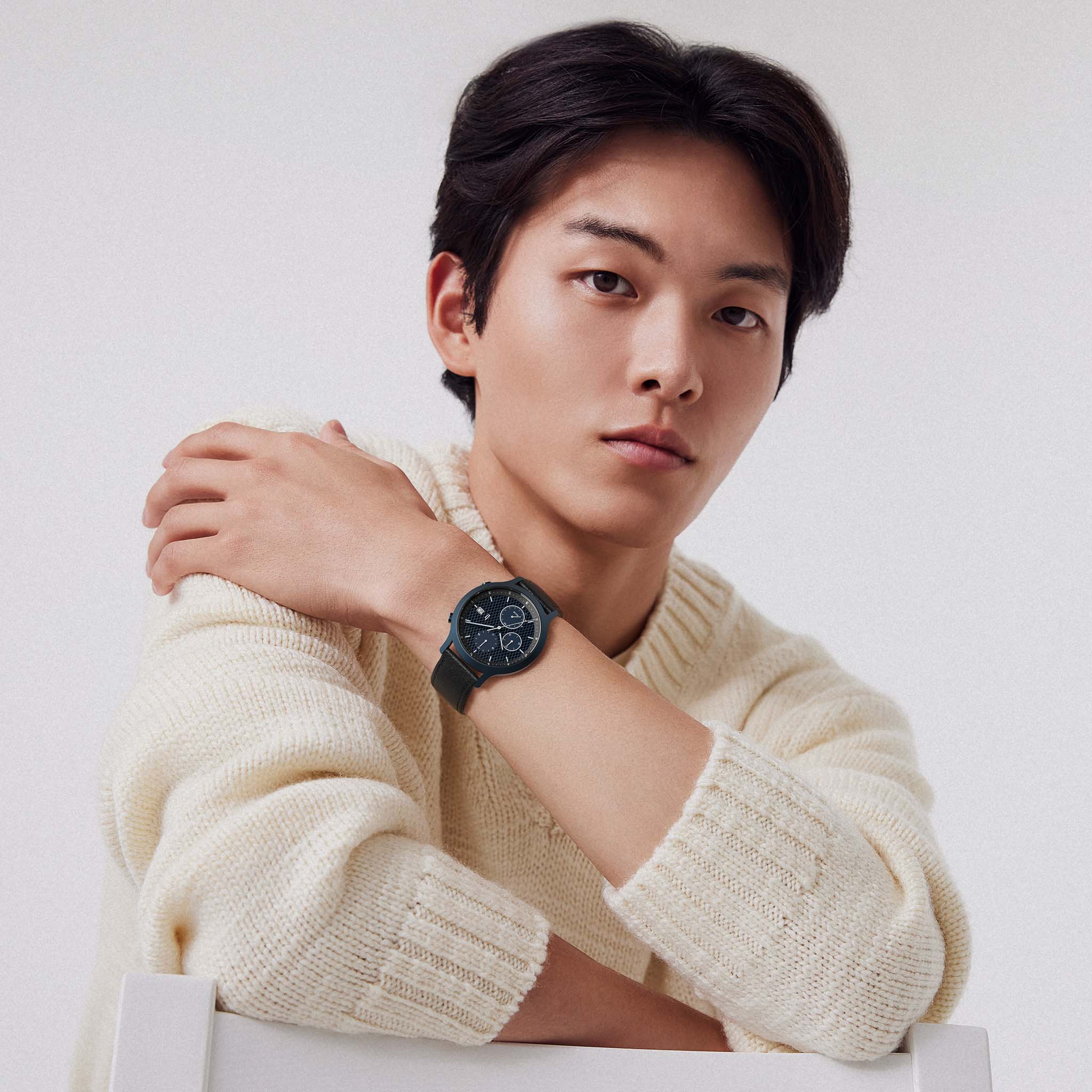 Man in cream sweater wearing a black chronograph wristwatch, seated against plain background