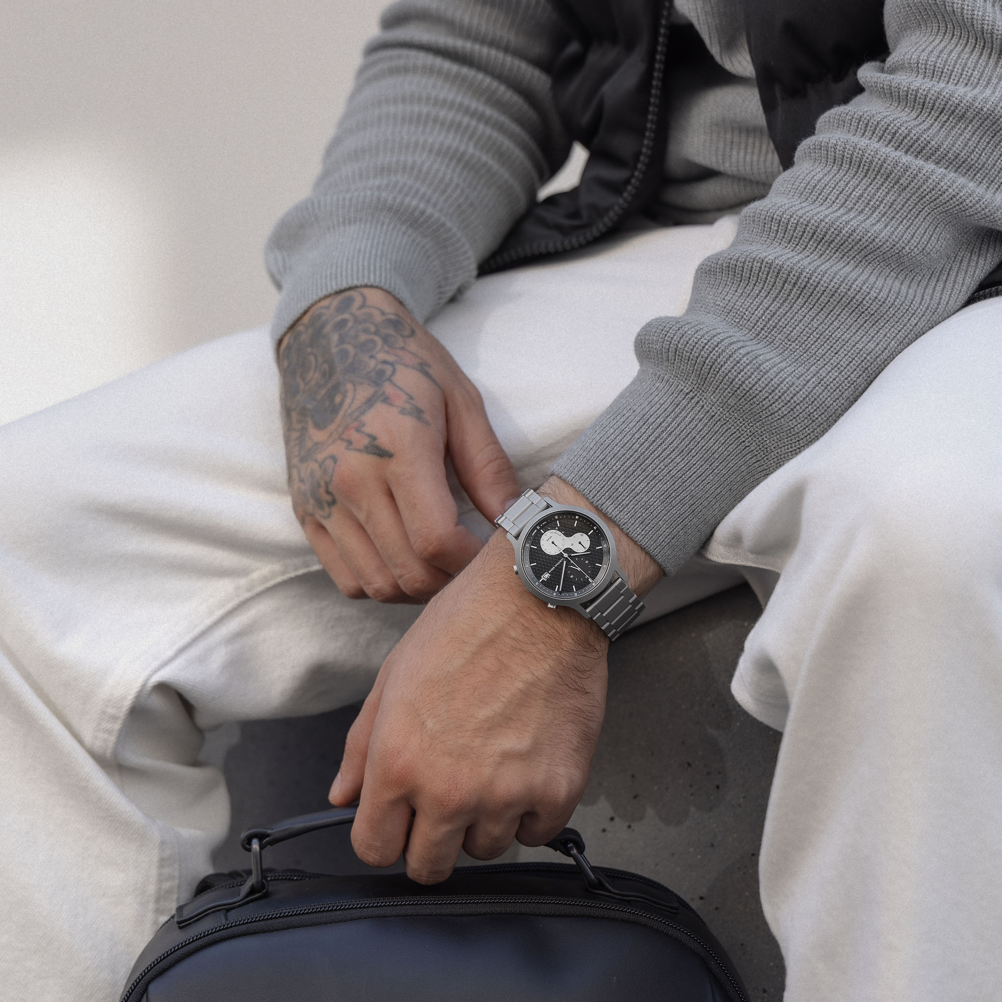 Man wearing a modern stainless steel wristwatch with black dial, holding a black bag.