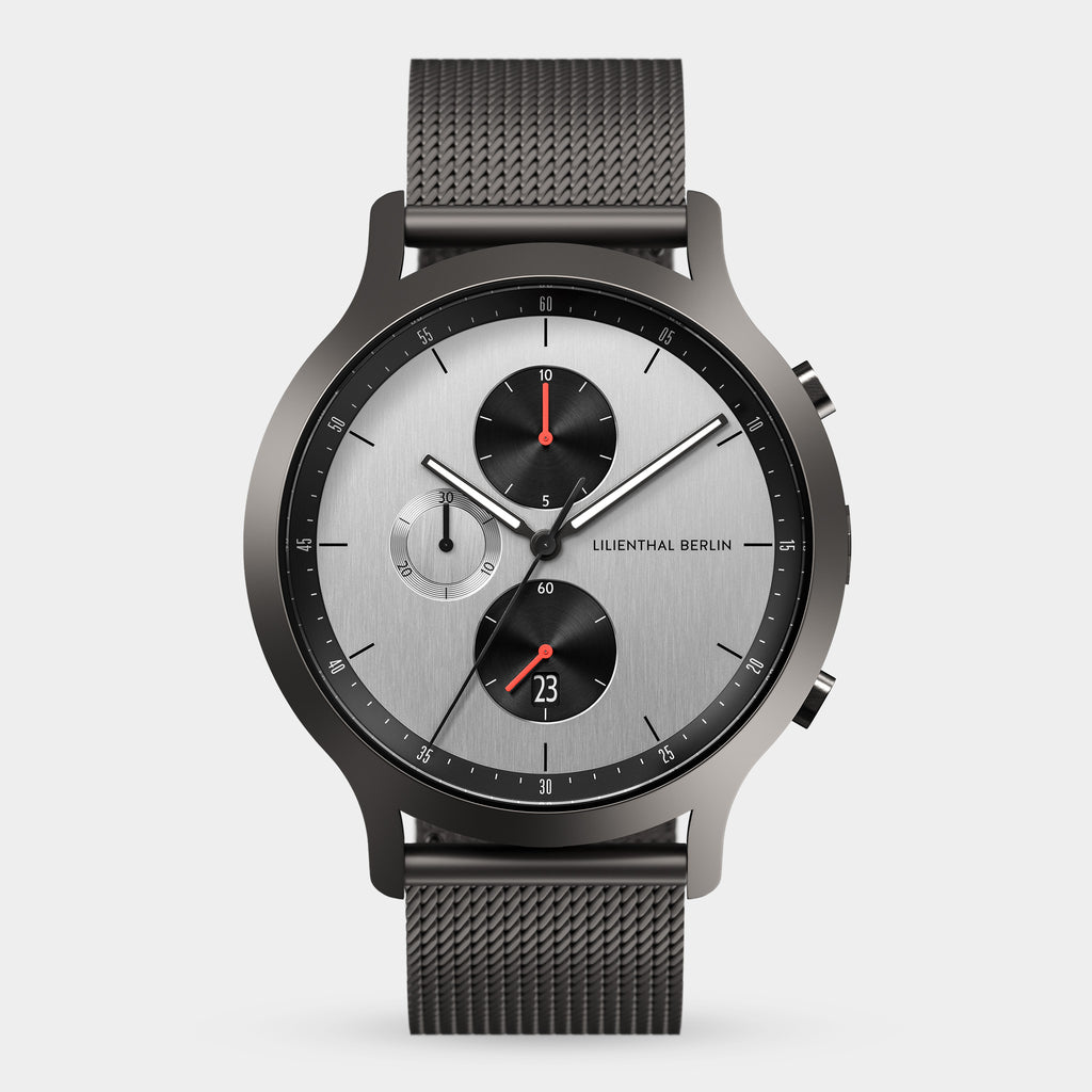 Minimalist silver chronograph watch with black mesh strap, three subdials, and red accents.