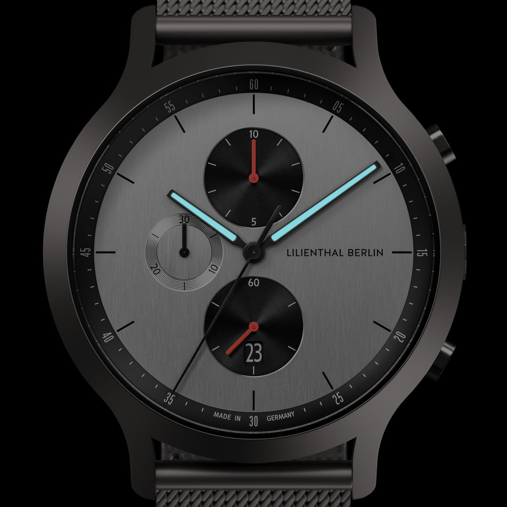 Minimalist black and silver chronograph watch with mesh strap, subdials, and blue hands