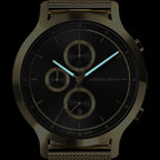 Close-up of a black chronograph watch with gold accents, mesh strap, and luminous hands.